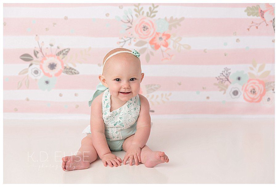 Smiling baby girl in blue outfit, sitting on the floor leaning forward with a big smile.